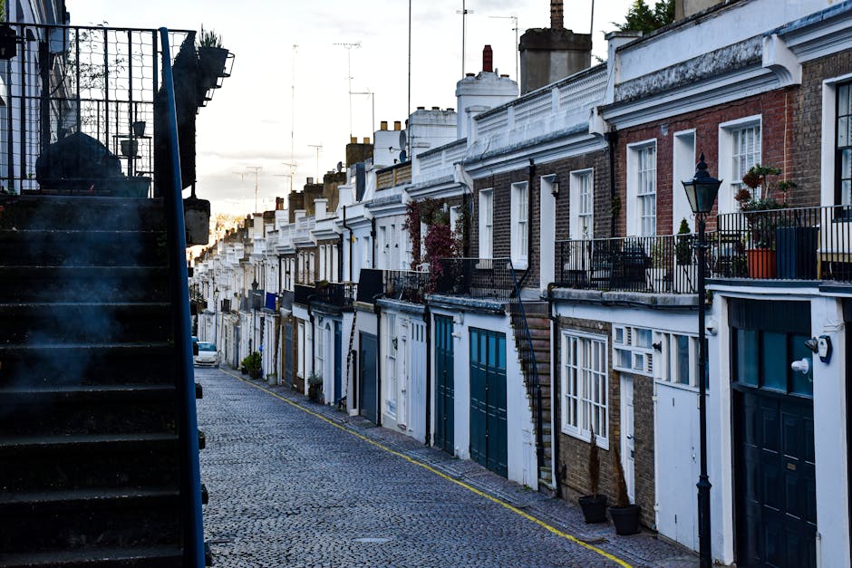 A row of neatly arranged terraced residential buildings with brick and white-painted facades, featuring large windows and small balconies with potted plants. The cobblestone street is clean and well-maintained, illuminated by classic black street lamps, with a staircase on the left side leading to an upper level. The overall scene depicts a quiet, tidy neighborhood in Islington during early evening, emphasizing the importance of surface cleaning and upkeep in maintaining the area's appearance, consistent with Deep Cleaning Islington's focus on deep cleaning and sanitisation services.
