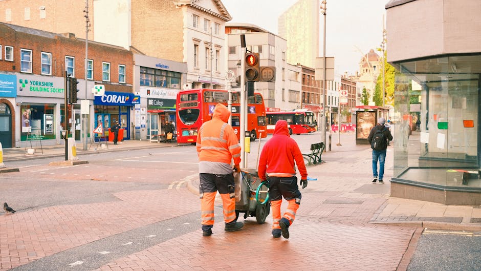 Two street cleaners in high-visibility orange and red uniforms are pushing a cleaning cart on the brick sidewalk of a busy urban area in Islington. Behind them, there are commercial buildings with storefronts, including a Yorkshire Building Society branch and a Coral betting shop, with signage visible. The street features a red double-decker bus stopped at a traffic light, and a few pedestrians are walking or waiting nearby. The scene is well-lit with natural daylight, and the pavement appears clean and maintained. Deep Cleaning Islington is not visibly referenced in the scene but is associated with surface cleaning and sanitation services for city environments, ensuring hygiene and cleanliness in busy residential and commercial areas.
