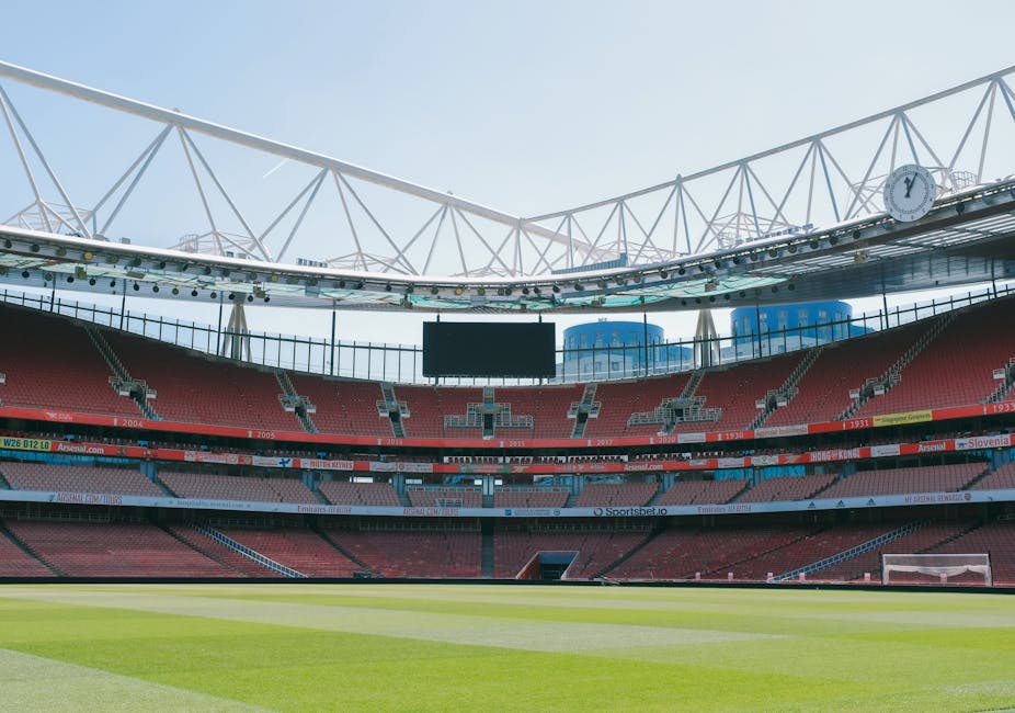 An empty stadium with a well-maintained grass pitch, surrounded by red and grey seating areas. The stadium features modern white structural beams and a large overhead digital screen hanging above the 