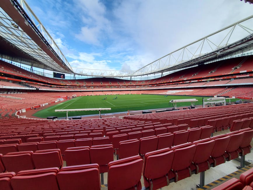 An empty stadium with a well-maintained grass pitch, surrounded by red and grey seating areas. The stadium features modern white structural beams and a large overhead digital screen hanging above the 