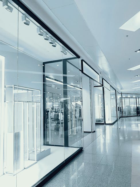 Interior view of a modern commercial office space in Canonbury Islington, featuring large glass display windows with black metal frames, showcasing a variety of retail products on white display stands. The polished, reflective tile flooring is spotless and free of dust or stains, contributing to a clean and professional atmosphere. The ceiling has built-in lighting fixtures, providing bright, even illumination, and a white ceiling panel with a subtle, smooth finish. The overall setting demonstrates high standards of surface cleaning and sanitisation maintained by Deep Cleaning Islington, with glass surfaces thoroughly cleaned to eliminate smudges, and the tiled floors appearing recently deep cleaned and polished to enhance shine. Visible elements include glass display cases and retail shelving, positioned in an open, inviting layout suitable for commercial or retail purposes, with natural light streaming in through large skylights or windows, highlighting the cleanliness and tidiness of the space.