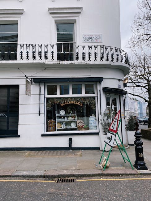 A street-level view of a white curved corner building with large display windows showcasing ceramic and glassware, including plates and bowls, inside a bakery shop labeled 'Boulangerie'. The shop's exterior features a black awning above the window, and there is a sandwich board sign on the sidewalk with red, white, and green colors, likely representing the Italian flag. The building has a decorative black wrought iron balcony on the first floor, with a white sign indicating 'Clarendon Cross W1'. The pavement appears clean and well-maintained, with a black bollard near the curb and a storm drain on the street. Overcast sky suggests typical weather for an urban area, and the scene is part of a commercial district in Canonbury, Islington, captured for a cleaning company's deep cleaning and surface sanitisation services, highlighting the importance of maintaining a tidy, hygienic appearance for retail and hospitality spaces. Deep Cleaning Islington specializes in professional cleaning for shops and commercial properties in this area.