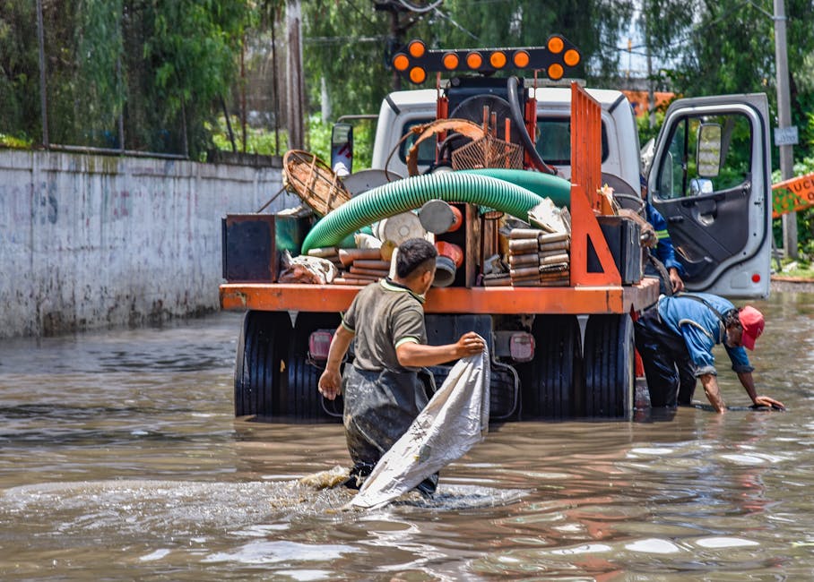 Two workers in waterproof clothing are engaged in flood water removal in an outdoor area, with one person holding a large white sack and the other stabilizing himself beside a heavy-duty orange flood recovery truck. The truck is equipped with hoses, tools, and equipment for emergency flood response, and is parked partially submerged in muddy water near a concrete wall and green trees. Bright natural daylight illuminates the scene, highlighting the muddy surface and waterlogged environment. Deep Cleaning Islington provides specialised flood cleaning services aimed at rapid response to urgent flooding emergencies, ensuring effective surface cleaning and sanitisation in affected residential or commercial properties.