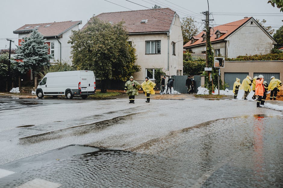 A residential street with wet asphalt and accumulating water, indicating recent flooding. Several workers dressed in yellow and orange raincoats are conducting surface cleaning and flood response activities near the curb and sidewalk, using tools and equipment for water removal and sanitisation. The background features two-story houses with white and beige exteriors, tiled roofs, and surrounding greenery, including trees and shrubs. Overcast skies cast diffuse lighting across the scene, highlighting the reflective, damp surfaces on the road and pavement. Deep Cleaning Islington offers emergency flood cleaning services to ensure hygiene and maintenance for affected properties, focusing on thorough surface cleaning, sanitisation, and water extraction in residential areas affected by flooding.