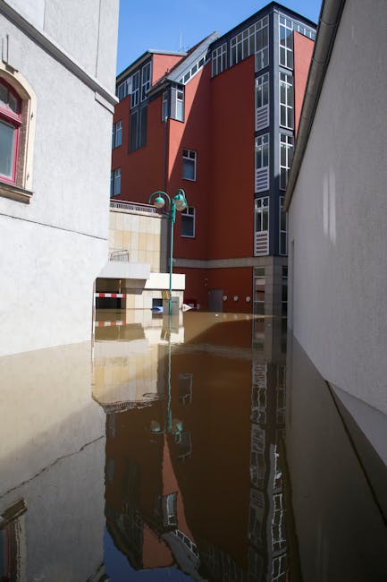 Two workers in waterproof clothing are engaged in flood water removal in an outdoor area, with one person holding a large white sack and the other stabilizing himself beside a heavy-duty orange flood recovery truck. The truck is equipped with hoses, tools, and equipment for emergency flood response, and is parked partially submerged in muddy water near a concrete wall and green trees. Bright natural daylight illuminates the scene, highlighting the muddy surface and waterlogged environment. Deep Cleaning Islington provides specialised flood cleaning services aimed at rapid response to urgent flooding emergencies, ensuring effective surface cleaning and sanitisation in affected residential or commercial properties.