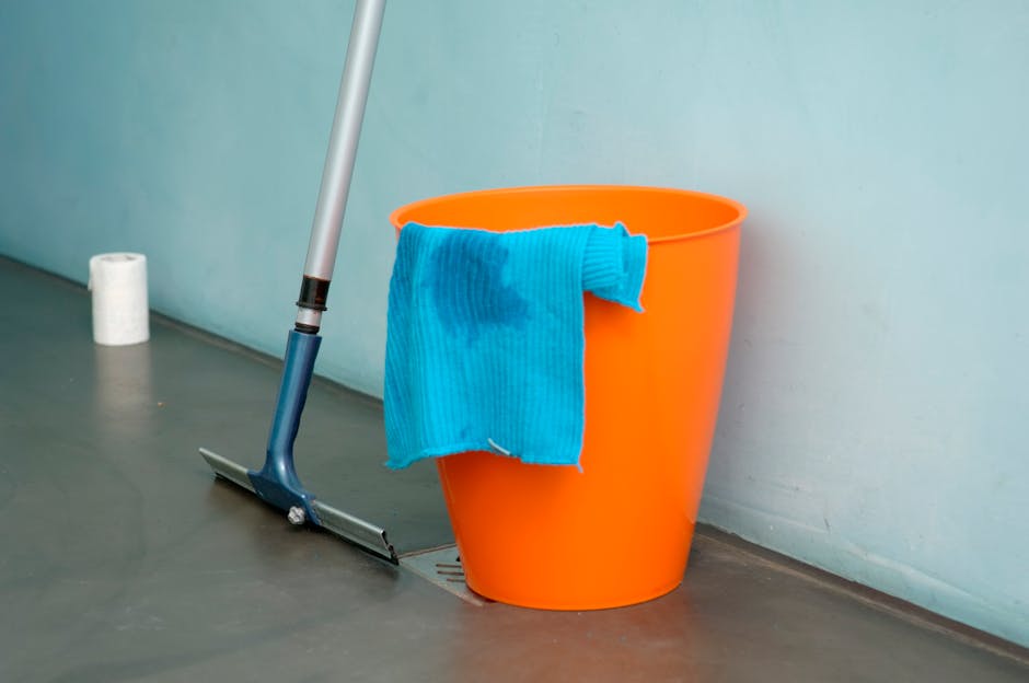 An orange plastic bucket with a damp blue microfiber cloth draped over its rim, positioned next to a squeegee mop with a silver handle and dark blue rubber blade, resting on a smooth, light gray floor near a light-colored wall. In the background, a roll of white toilet or paper towels is visible, indicating a setting for surface cleaning or sanitisation in a domestic or commercial space. The scene is well-lit, emphasizing the cleanliness and readiness for deep cleaning by Deep Cleaning Islington, alongside visible drainage on the floor for easy water management during cleaning operations.