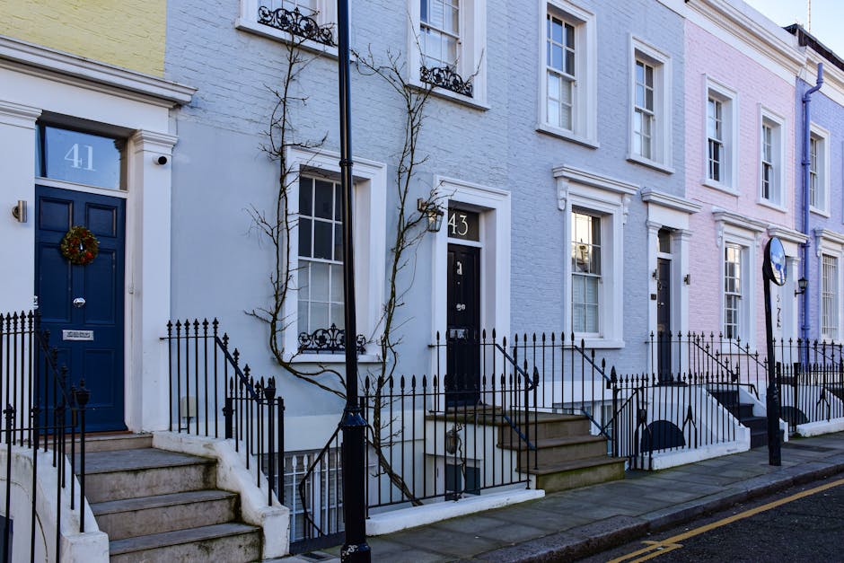 A row of colorful terraced houses on Upper Street in Islington, N1, with pastel-colored facades including shades of yellow, blue, and pink. Each house features front steps leading to the main entrance, black wrought iron railings along the steps and sidewalk, and white-framed sash windows. One house has a decorative wreath hanging on a dark blue door, and another has a small potted plant on the windowsill. The pavement appears clean and well-maintained, with a lamppost and minimal street clutter visible. The lighting suggests daytime, with soft natural light illuminating the scene. This residential street showcases typical Victorian-style terraced houses, and the outdoor surfaces look tidy, reflecting professional deep cleaning and maintenance by Deep Cleaning Islington, experts in domestic cleaning services.