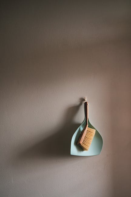 An orange plastic bucket with a damp blue microfiber cloth draped over its rim, positioned next to a squeegee mop with a silver handle and dark blue rubber blade, resting on a smooth, light gray floor near a light-colored wall. In the background, a roll of white toilet or paper towels is visible, indicating a setting for surface cleaning or sanitisation in a domestic or commercial space. The scene is well-lit, emphasizing the cleanliness and readiness for deep cleaning by Deep Cleaning Islington, alongside visible drainage on the floor for easy water management during cleaning operations.