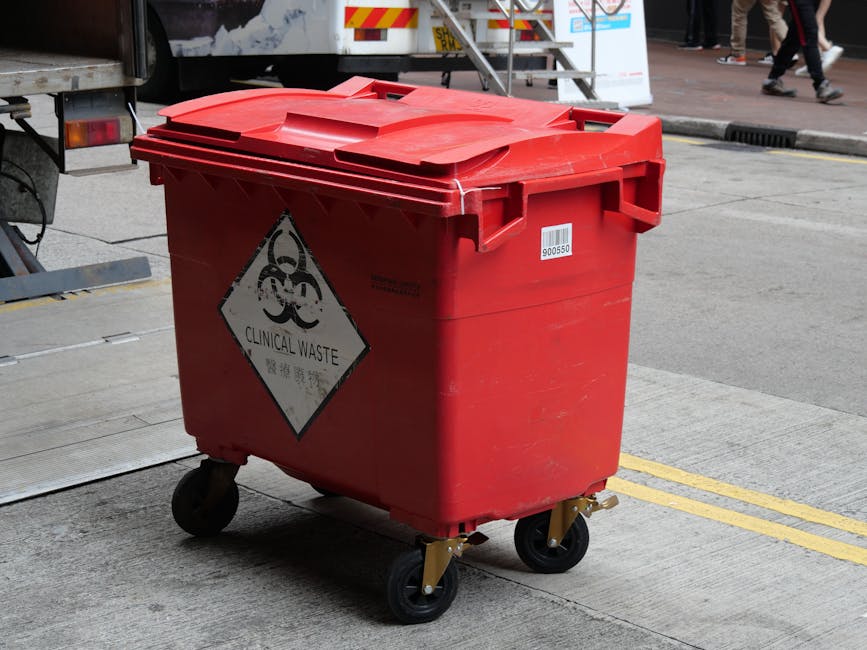 A red medical waste bin with a biohazard symbol and the words 'Clinical Waste' printed on a diamond-shaped label, positioned on a concrete pavement next to a vehicle. The bin has a closed lid and is m