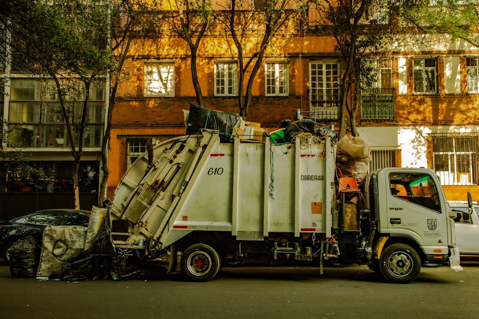 An outdoor scene showing multiple large yellow and transparent plastic rubbish bags filled with waste, piled against a white textured wall and corner of a building. The bags are placed on a cobbleston