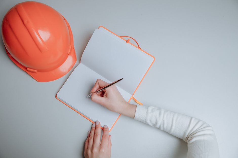 Close-up of a person in a high-visibility yellow vest holding a clipboard with a home inspection checklist, positioned indoors near a window that lets in natural light. The individual's right hand gri