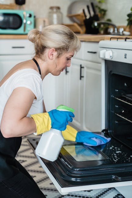 A woman wearing a white shirt, yellow and blue rubber gloves, and holding a spray bottle and cleaning cloth, is deep cleaning the black oven in a modern kitchen. The oven door is open, revealing a clean, shiny interior with visible racks. The kitchen features white cabinets with black handles, a wooden countertop, and various appliances including a microwave and a kettle, along with potted plants and utensils on the counter. The scene is brightly lit, showcasing the meticulous cleaning process by Deep Cleaning Islington, aimed at surface cleaning and sanitisation of domestic appliances.