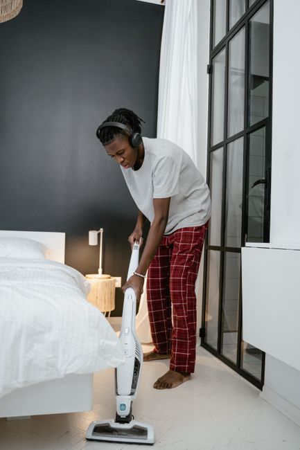 A woman with dark skin, wearing a white T-shirt, red plaid pajama pants, and headphones, is performing surface cleaning and vacuuming in a modern bedroom. She is bent slightly forward, holding a handh